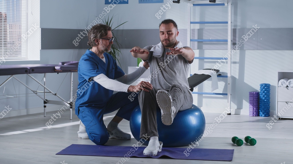 Middle Aged Therapist Helping Patient To Balance On Fit Ball Middle Aged Therapist Helping Patient To Balance On Fit Ball