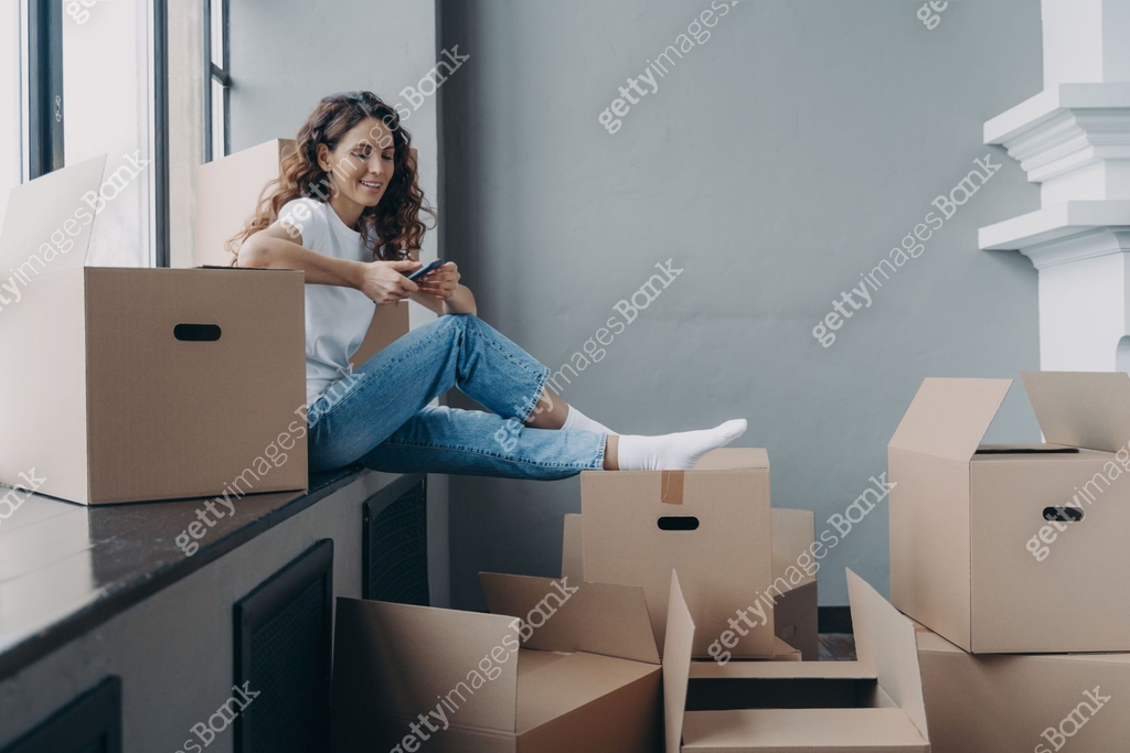 Cheerful european woman is sitting on windowsill packing boxes and ...