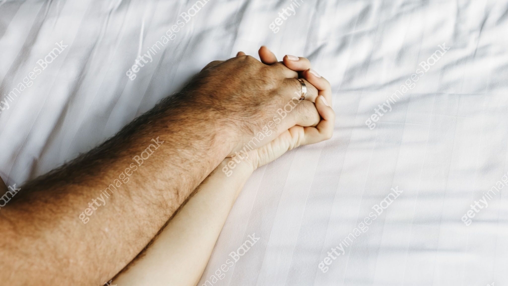 A close-up of a couple's hands intertwined on the bed, capturing a ...